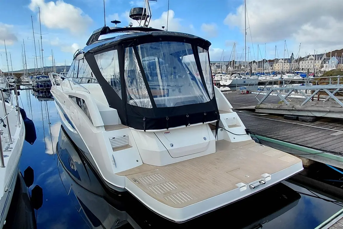 A luxury motorboat docked in a marina, with clear reflections and nearby yachts, framed by a cloudy blue sky.